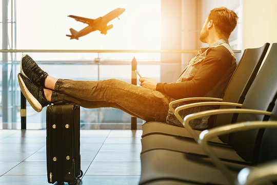 Man sitting in an airport lounge with feet on his suitcase, using his phone while an airplane takes off, highlighting the convenience of staying connected with an eSIM for travel.