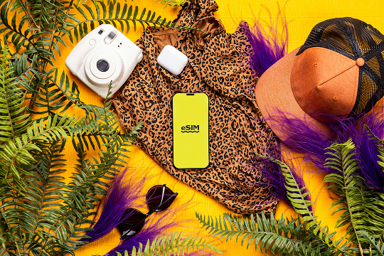 A stylish travel flat lay featuring a smartphone displaying 'eSIM', surrounded by vacation essentials like sunglasses, a hat, a camera, and tropical leaves, representing Montenegro travel connectivity.